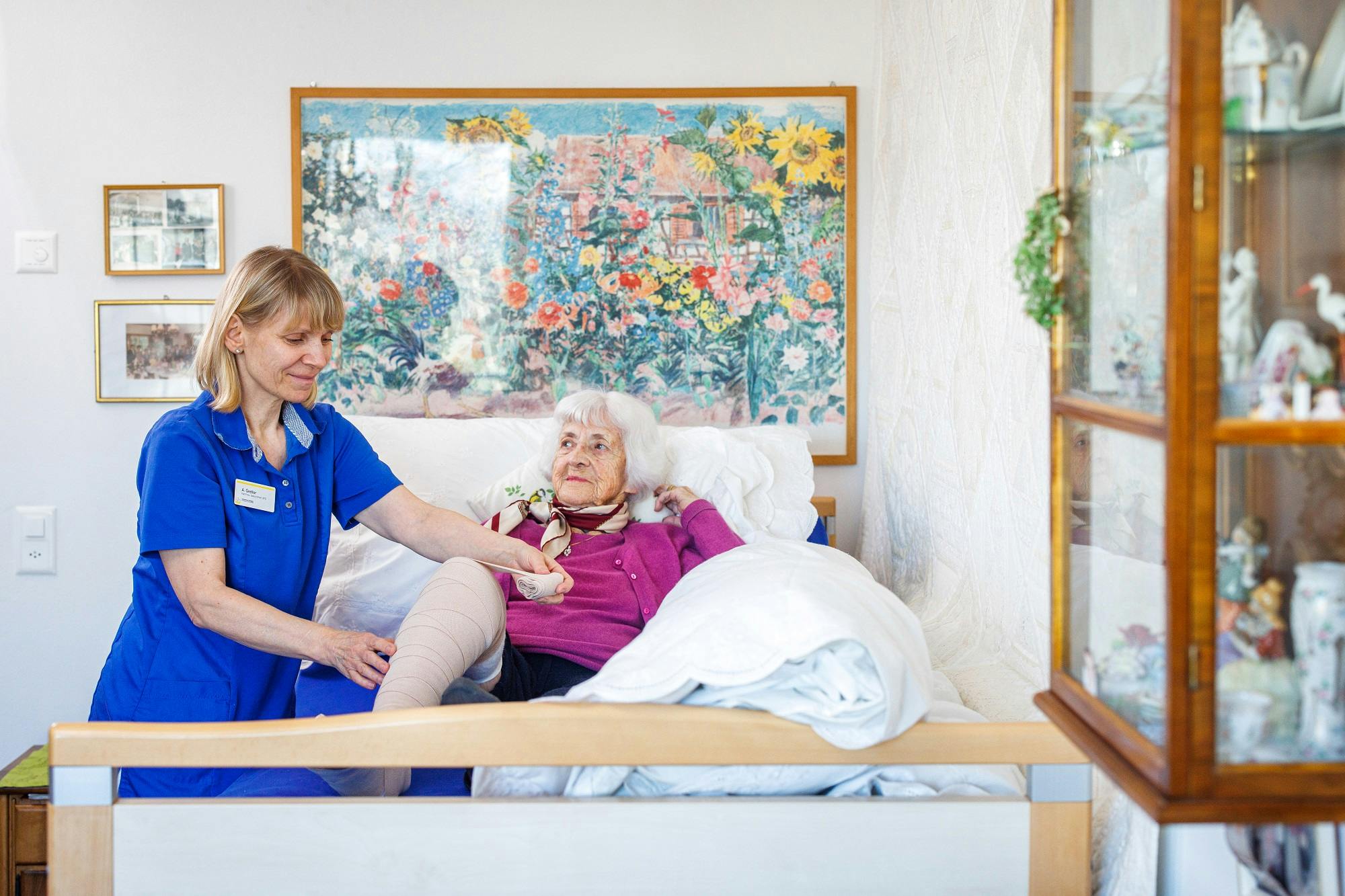 Pflegekraft betreut ältere Dame im Bett vor Blumenbild.