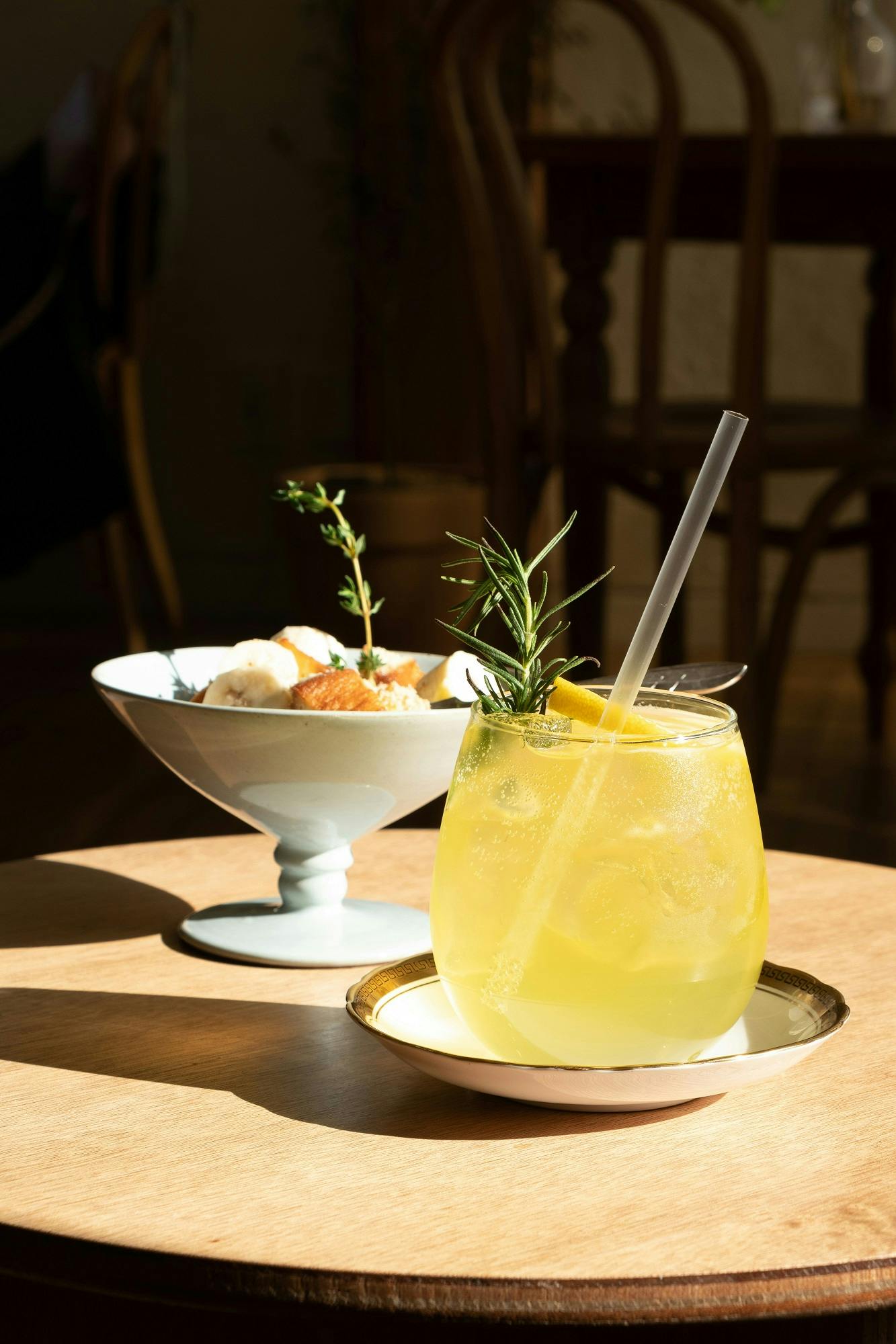 Cocktail mit Rosmarin und Eiswürfeln auf einem Teller, daneben eine Schale mit Eiscreme, im Hintergrund eine sonnendurchflutetes Café.